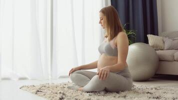 Pregnant woman in sportswear sitting on the carpet in the lotus position. Health care during pregnancy. Yoga for pregnant women video