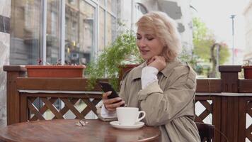 vrouw toepassingen de telefoon met een kop van koffie Bij een tafel Aan de terras. een vrouw in een loopgraaf jas en wit blouse picks omhoog een wit koffie kop en zet het terug Aan de tafel video