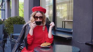 elegante caffè. il bionda bevande caffè con un' brioche. moderno nero terrazza di il ristorante. giovane donna nel occhiali da sole e berretto video