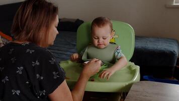 mamá alimentación bebé chico en alto silla desde cuchara, familia simpático atmósfera video