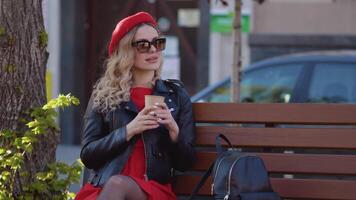 jong mooi vrouw in een rood baret en jurk, zwart bril en een vlecht is zittend Aan een park bank met kop van koffie. lunch breken Bij werk in de midden- van de werken dag video