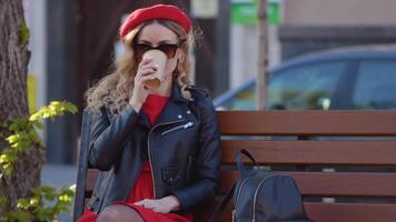 Young beautiful woman in a red beret and dress, black glasses and a braid is sitting on a park bench with cup of coffee. Lunch break at work in the middle of the working day video