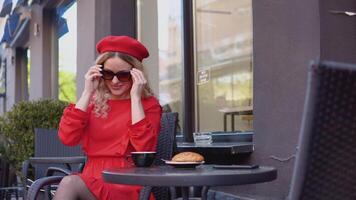 jovem mulher sorridente e vestindo à moda oculos escuros. mulher dentro uma vermelho vestir e boina sentado com uma copo do café e uma croissant em uma mesa em a rua video