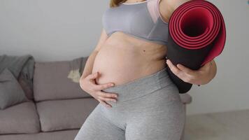 gravid kvinna i sportkläder stående med kondition matta i händer på Hem i levande rum. hälsa och sporter vård under graviditet. närbild se av de mage och matta video