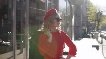 Stylish young blonde woman in a red dress and black braid stands on a cozy street in the morning sun video