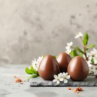 AI generated Three glossy chocolate eggs lie on a slate with white blossoms against a neutral grey backdrop. perfect for springtime Easter promotions or minimalist culinary presentations. photo