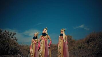 Three women in traditional dresses and headpieces holding hands in a barren landscape at dusk. video