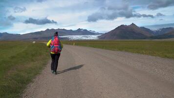 weiblich Tourist mit Rucksack geht durch Schmutz Straße auf Hintergrund von Berge und Gletscher im Island. Freiheit und Reise Konzept. 4k video