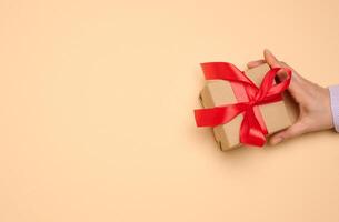 A woman's hand holds a gift box wrapped in a red silk ribbon on a beige background photo
