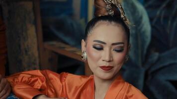 elegante mujer con tradicional peinado y constituir, vistiendo naranja atuendo, posando con un sereno expresión. video