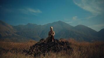 man in traditional dress standing on rocky terrain with mountainous backdrop. video