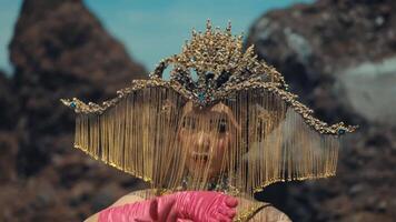 Surreal portrait of a person with an avant-garde headpiece and pink gloves, posing against a rocky background. video