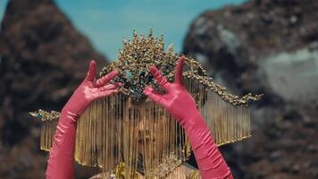 Woman in elaborate golden headpiece and pink gloves posing against a rocky background. video