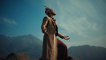 persoon in wijnoogst kostuum met armen uitgestrekt tegen bergachtig backdrop onder blauw lucht. video