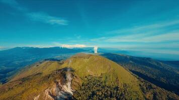 Antenne Aussicht von ein üppig Grün Berg Landschaft mit ein entfernt Wasserfall unter ein klar Blau Himmel. video
