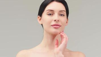 Close up beauty portrait of young woman with smooth healthy skin, she gently touches her face with her fingers on light grey background and smiles video