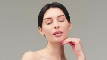 Close up beauty portrait of young woman with smooth healthy skin, she gently touches her face with her fingers on light grey background and smiles video