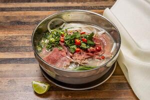 Hanoi rare Beef Pho topping with onion, noodles, green onion, red chilli, lemon and coriander served in bowl isolated on wooden table top view of hong kong fast food photo