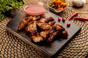 GRILLED CHICKEN WINGS with sauce and pickle served in a wooden cutting board isolated on wooden table background side view of appetizer photo