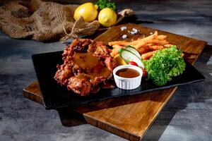 Crispy Boneless Chicken Steak in a black dish with french fries, sauce and lime top view on dark background photo