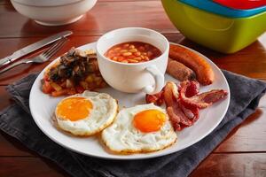 full american breakfast with sausage, sunny side up eggs and beans served in a dish isolated on wooden background side view photo