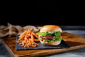 Delicious beef burgers with tomato, cheese, ketchup and french fries on wooden cutting board side view on dark background photo