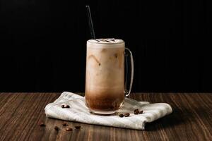 Iced mocha coffee with cream in a tall glass and coffee beans, portafilter, tamper and milk jug on dark wooden background. Cold summer drink. photo