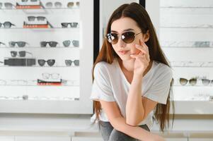 Portrait of a young woman shopping, standing in store and trying sunglasses near a mirror. photo