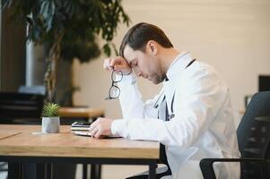 estresado masculino médico se sentó a su escritorio. medio adulto masculino médico trabajando largo horas. sobrecargado de trabajo médico en su oficina. no incluso doctores son eximir desde agotamiento . foto