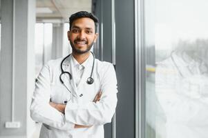 Portrait of male indian doctor wearing white coat having open door on clinic corridor as background photo