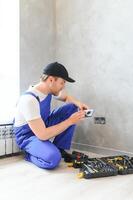 Confident professional electrician in uniform using screwdriver while replacing a socket in apartment after renovation work photo