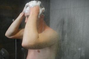 hermoso joven hombre tomando un ducha en pie debajo que cae agua gotas Lavado desnudo cuerpo y cabeza en moderno baño a hogar. masculino modelo foto