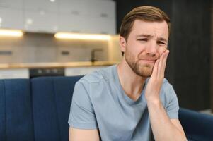 Toothache. Unhappy upset caucasian sad man sits on the couch at home, holds his hand near his cheek, grimaces, has an acute toothache, needs a consultation with a dentist doctor photo