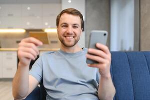 joven hombre sentado a hogar pago para comida en línea con teléfono inteligente y crédito tarjeta foto