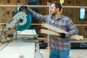 A carpenter works on woodworking the machine tool. Man collects furniture boxes. Saws furniture details with a circular saw. Process of sawing parts in parts. Against the background of the workshop. photo