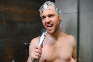 atractivo joven alegre hombre canto mientras Lavado en el ducha, vistiendo ducha gorra y participación ducha cabeza foto