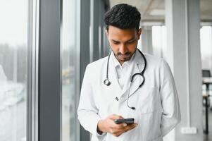 Portrait of male indian doctor wearing white coat having open door on clinic corridor as background photo