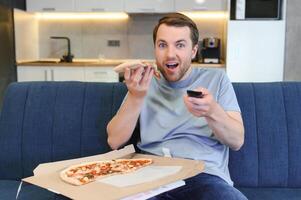 caucásico hombre descansando en sofá, comiendo Pizza y acecho televisión. concepto de moderno exitoso del hombre estilo de vida y relajarse después trabajar. cómodo estudio departamento. foto