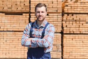 retrato de un hermoso trabajador elegir el mejor de madera tableros carpintero en pie siguiente a un grande apilar de madera barras en un depósito. foto