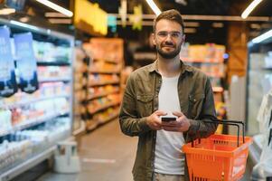 texto rápido durante las compras. apuesto joven sosteniendo un teléfono móvil y sonriendo mientras está de pie en una tienda de alimentos foto