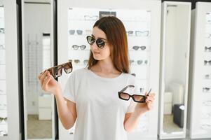 Portrait of a young woman shopping, standing in store and trying sunglasses near a mirror. photo