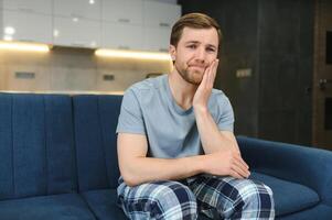 frustrado joven hombre conmovedor su mejilla mientras sentado en el sofá a hogar foto
