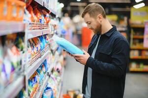 Young man in the supermarket in the household chemicals department. Large selection of products. A brunette in a glasses and a beard in a beige coat. photo