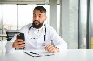 retrato de Arábica médico hermoso joven hombre en ropa de trabajo posando a moderno clínica, sentado a trabajo de Escritorio foto