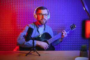 un joven elegante chico con un barba vistiendo casual o música profesor jugando guitarra en frente de teléfono inteligente cámara. en línea guitarra formación o vocal lección foto