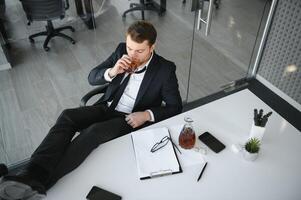 Fatigue young businessman sitting at workplace and holding whiskey bottle, drinking alcohol, bankruptcy concept. photo