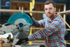 A carpenter works on woodworking the machine tool. Man collects furniture boxes. Saws furniture details with a circular saw. Process of sawing parts in parts. Against the background of the workshop. photo