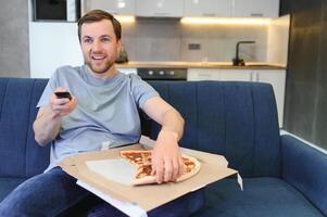 contento joven hombre Bebiendo cerveza y comiendo Pizza cuando acecho juego en televisión a hogar foto