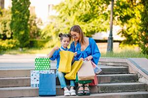 Beautiful mom and her cute little daughter are holding shopping bags, looking at camera and smiling while standing outdoors. Shopping concept. photo