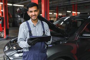 mecánico hombre mecánico gerente trabajador utilizando un ordenador portátil computadora comprobación coche en taller a auto coche reparar Servicio centro. ingeniero joven hombre mirando a inspección vehículo detalles debajo coche capucha. foto